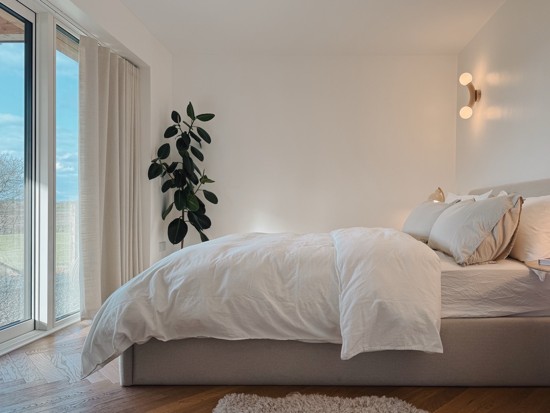 A bed with white linen sits beside glazed doors onto a garden, a potted plant in the corner and wall lights above the headboard.