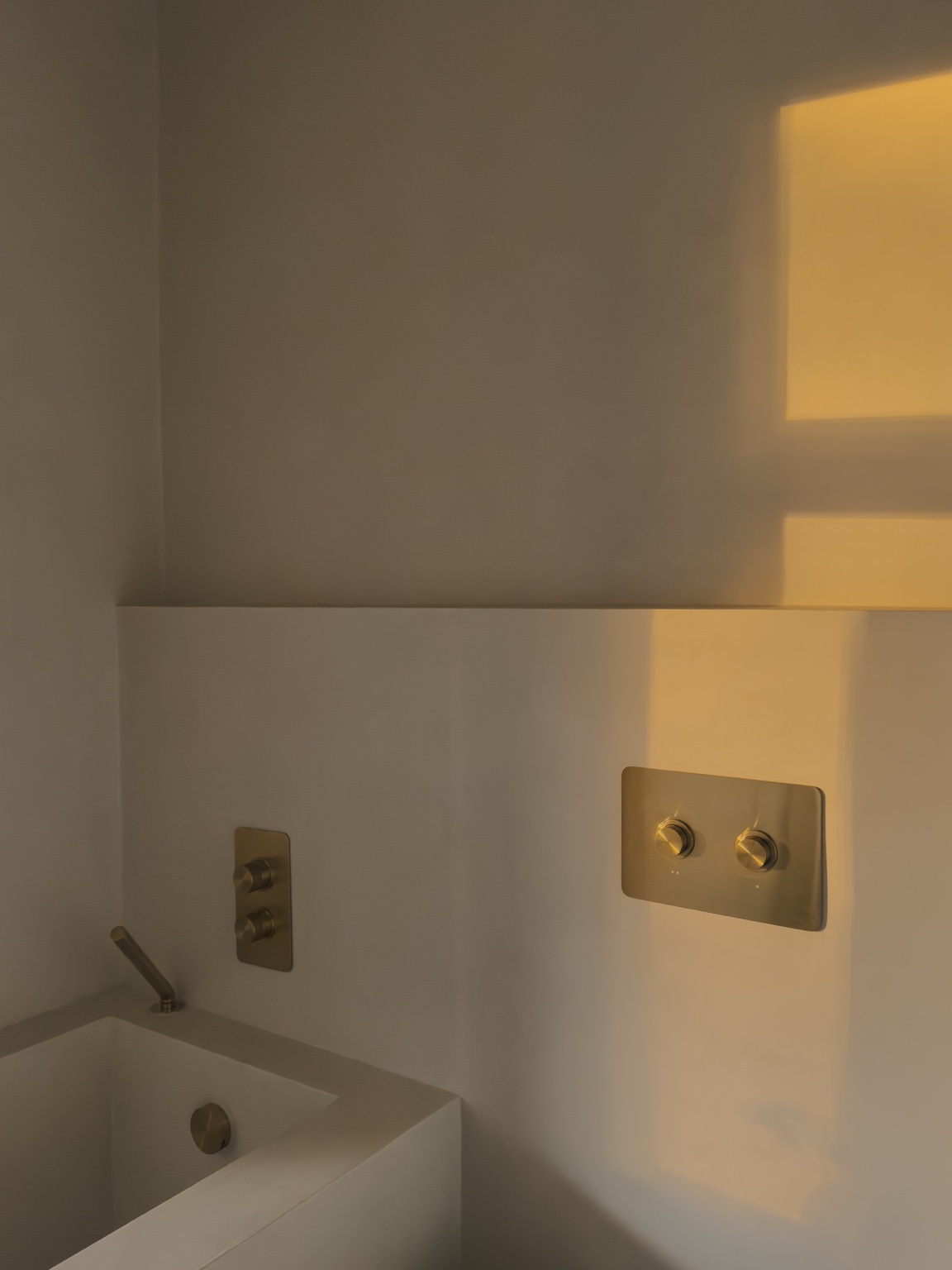 Morning light falls across a bathtub with brushed brass fittings and dimmer switches set into a plastered wall.