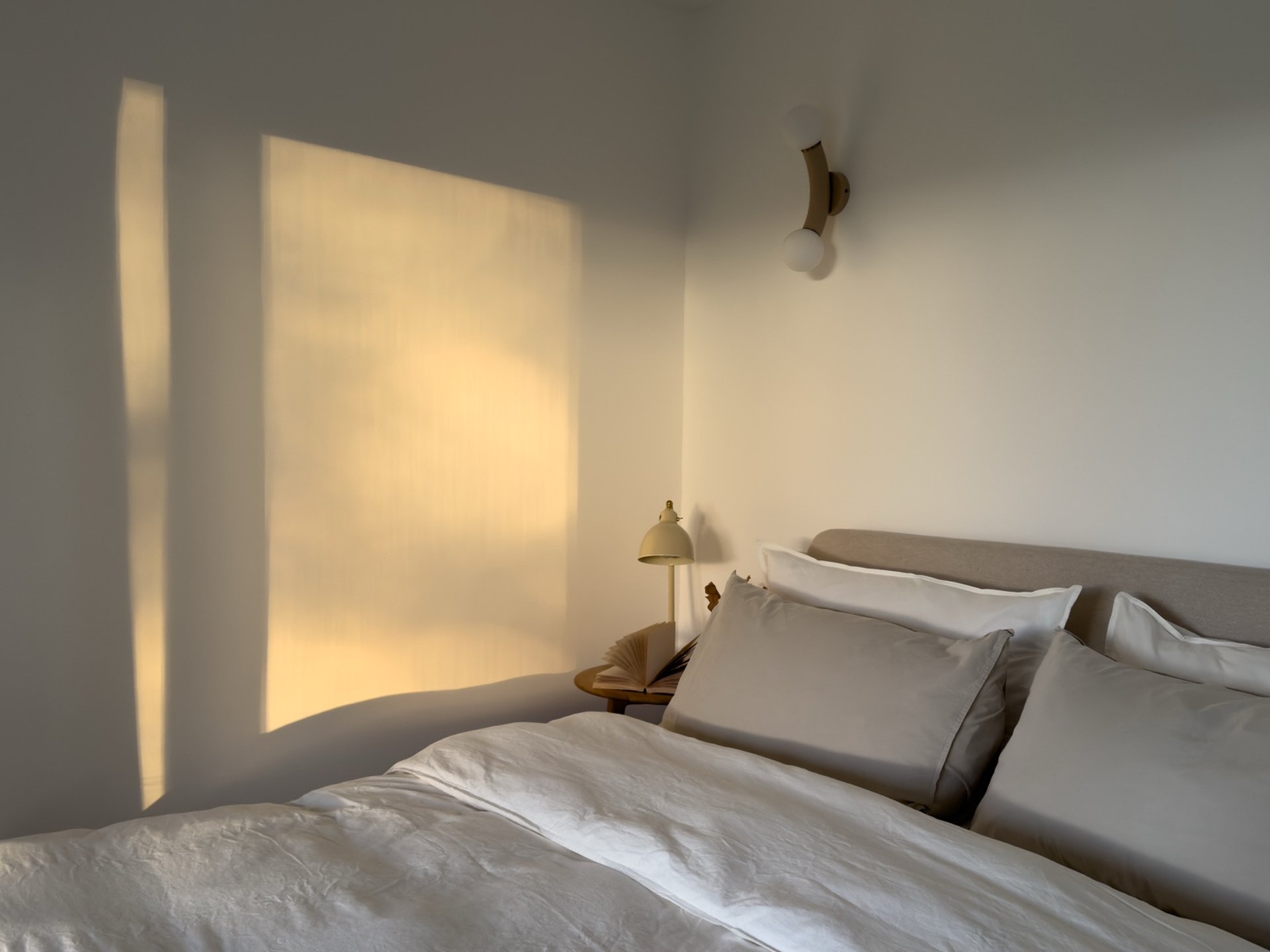 Evening light streams across a corner of a bedroom, a bed with white linen and a small bedside lamp in the foreground.