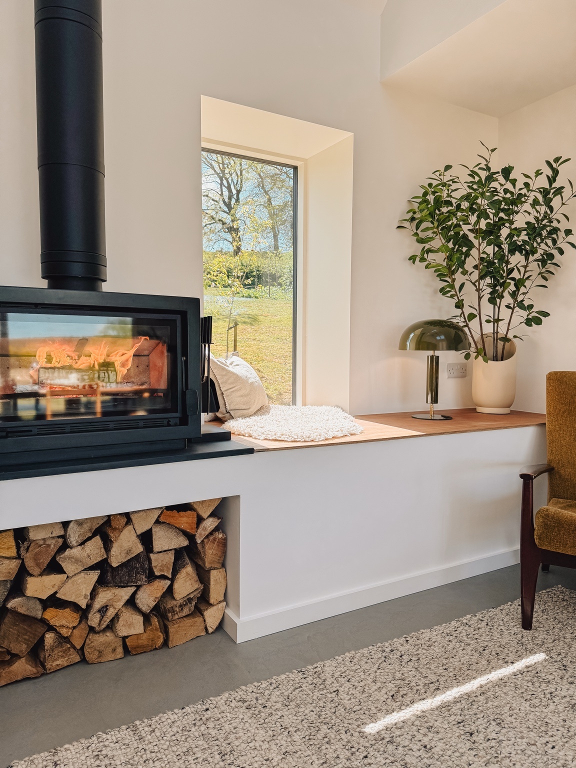 A wood burning stove is alight above a neat stack of logs, a window seat with a cushion and a potted plant beside an armchair.