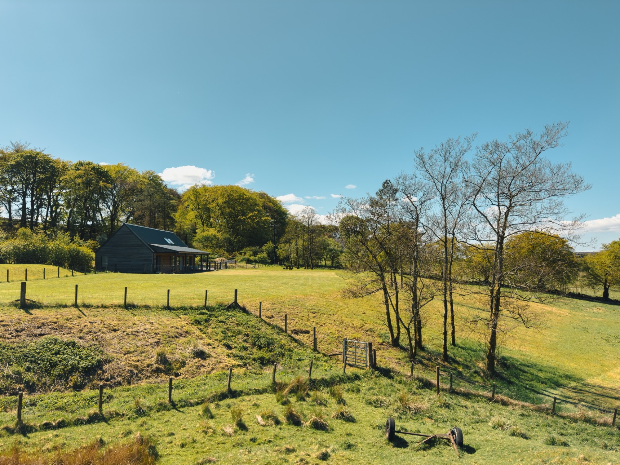 A dark timber house with a low-pitched roof sits in an open field bordered by post and wire fencing, trees framing the scene.