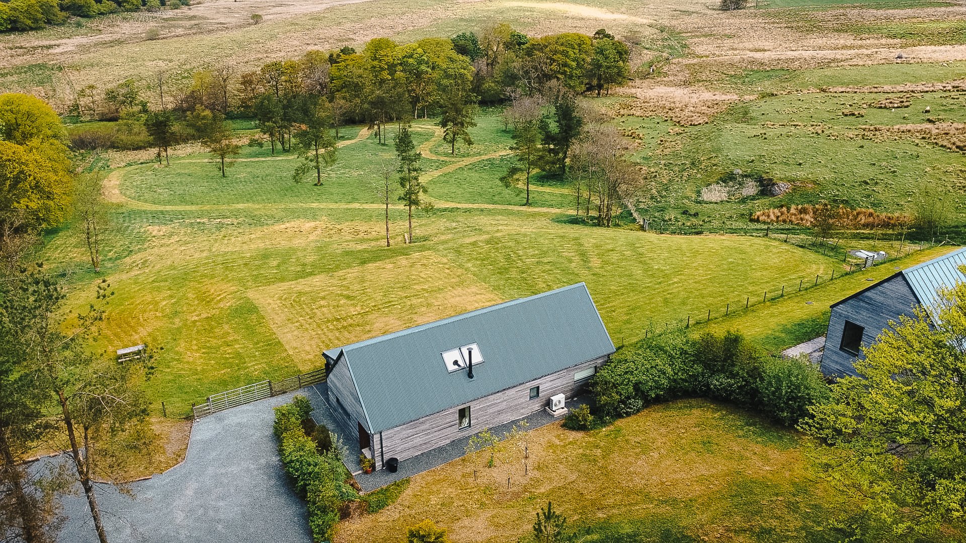 An aerial view shows a dark timber house with a pitched roof set in a mown lawn, surrounded by trees and open fields.