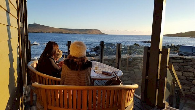 Two people on Fjarå's deck in the evening sun, looking over Breiwick Bay to Bressay.