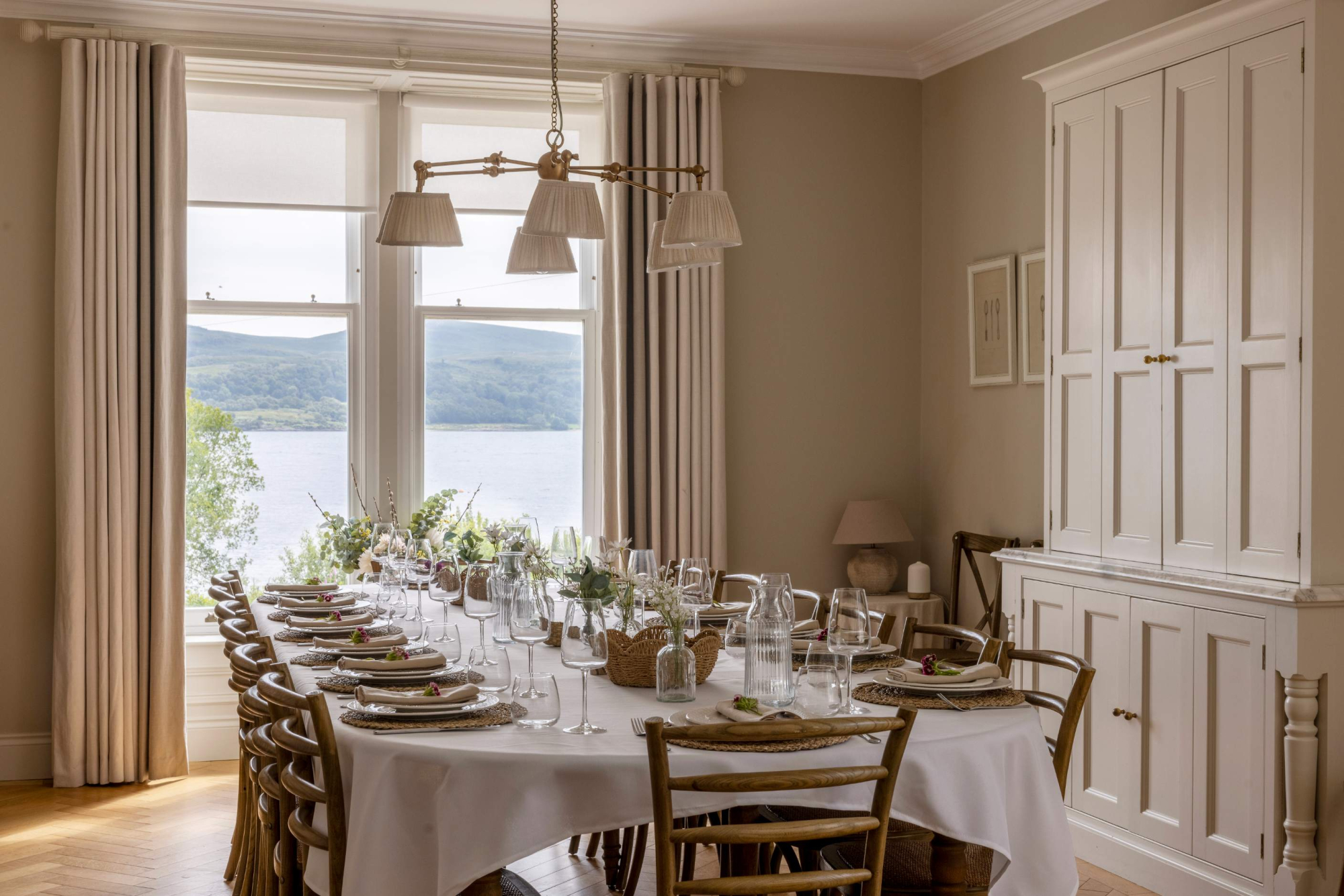 A long dining table set for a group stands beneath brass pendant lights, a loch and hills visible through the window and a panelled cabinet against the wall.