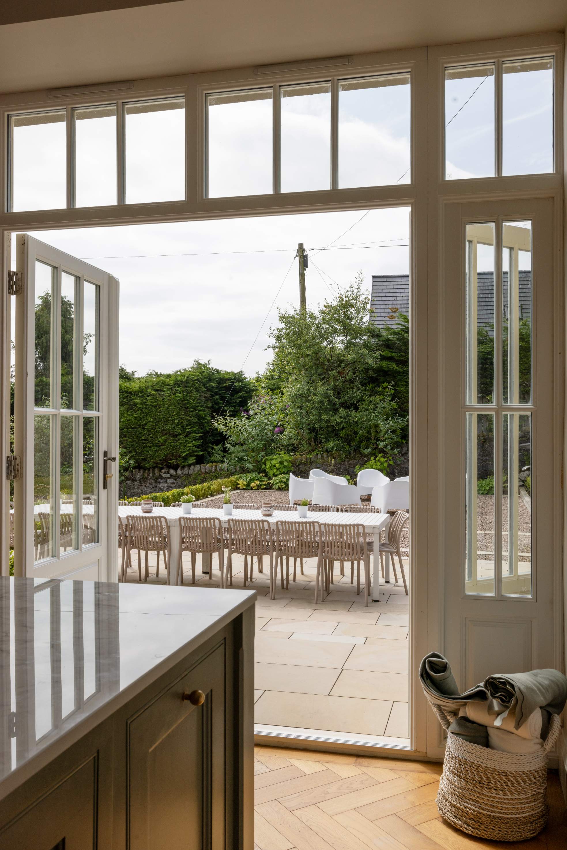 Open French doors lead from a marble-topped kitchen to a paved terrace set with a dining table, a woven basket of folded cloths beside the door.