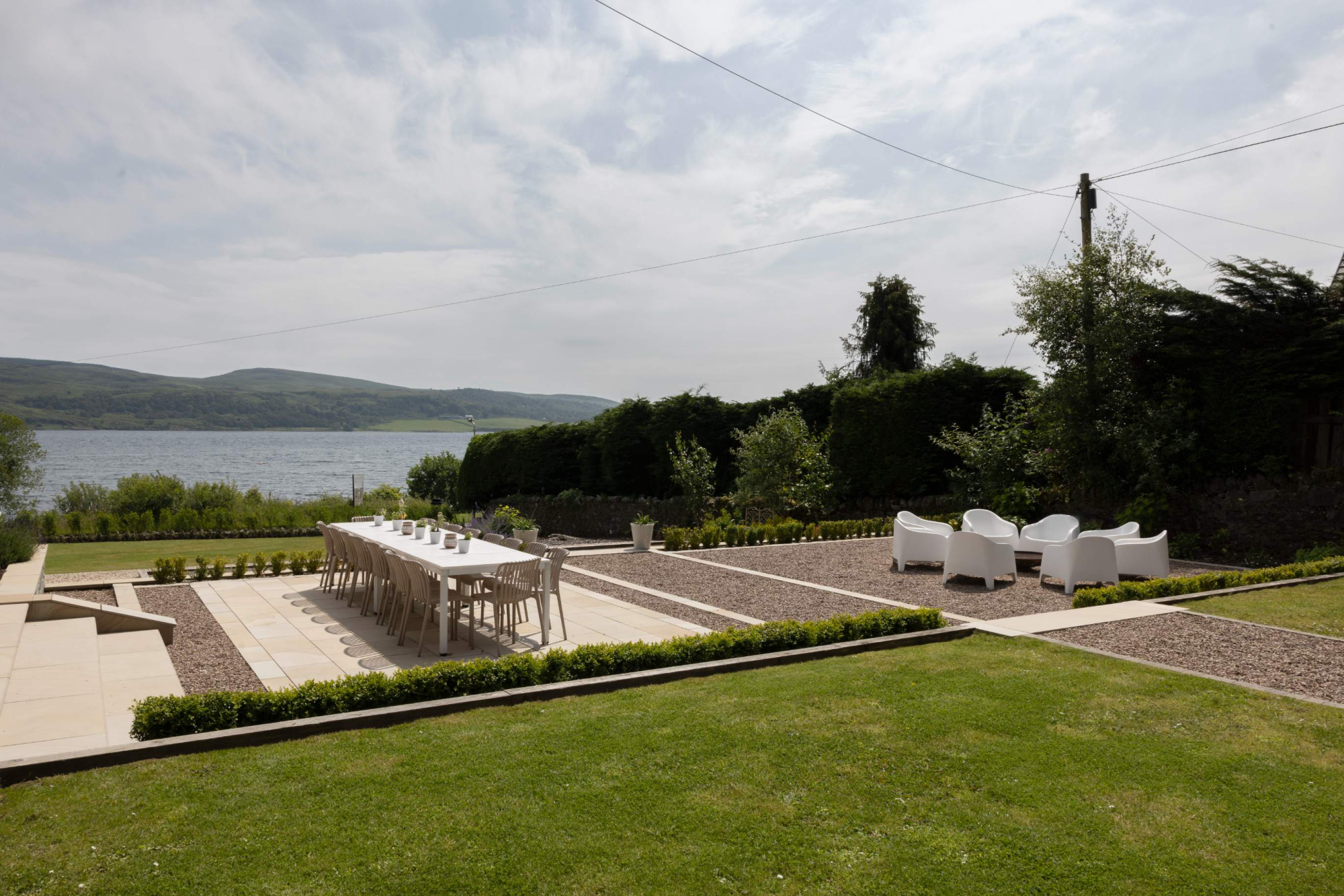 An elevated view shows a gravel patio with a dining table and white outdoor seating beside a lawn, a loch and hills stretching into the distance.