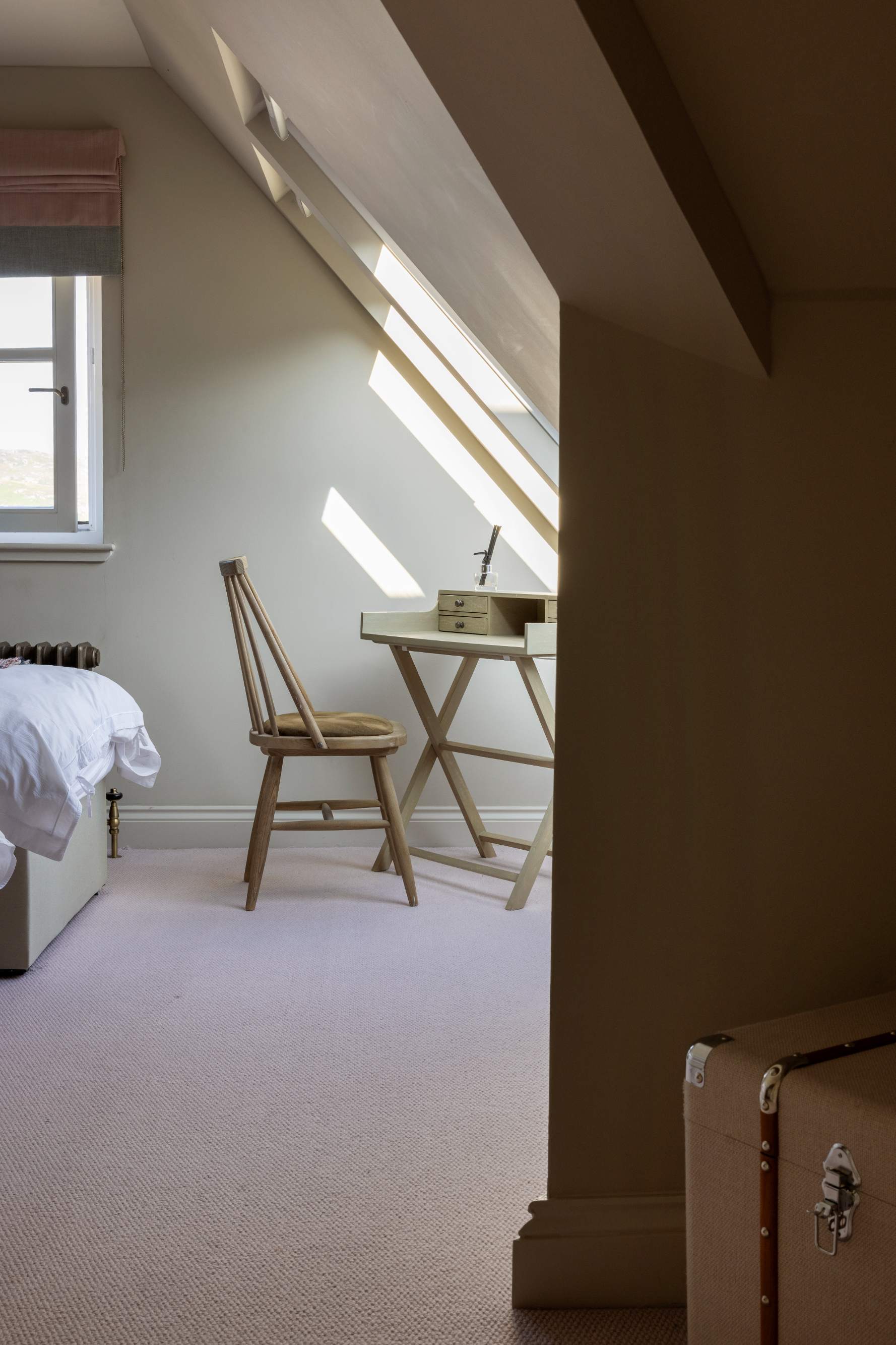 A wooden desk and chair sit beneath a sloped skylight in the corner of a bedroom, a vintage travel trunk and the edge of a bed nearby.
