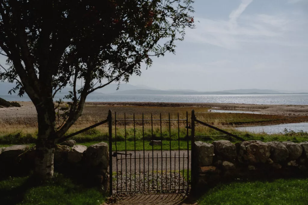 An iron gate in a stone wall opens onto a view of a beach and hills beneath a tree.