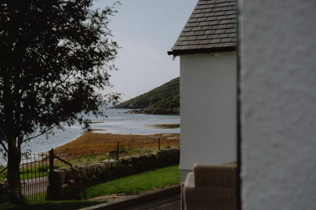 The white gable end of a cottage overlooks a sea loch, a stone wall and grass in the foreground.