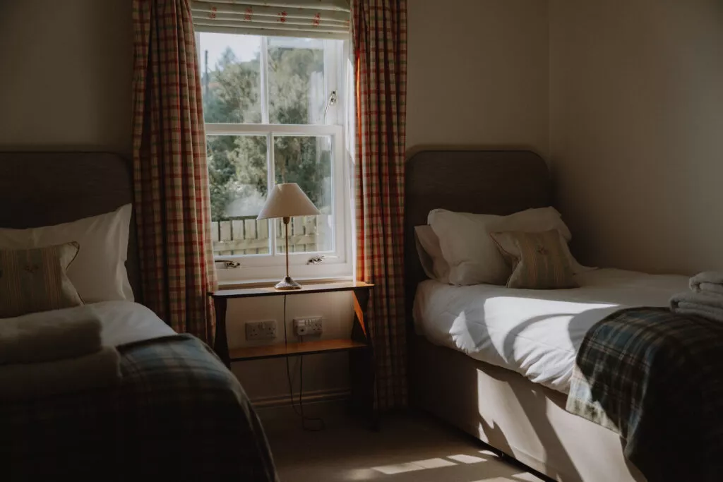 Twin beds with tartan headboards and plaid curtains flank a small table with a lamp, a folded towel resting on one bed.