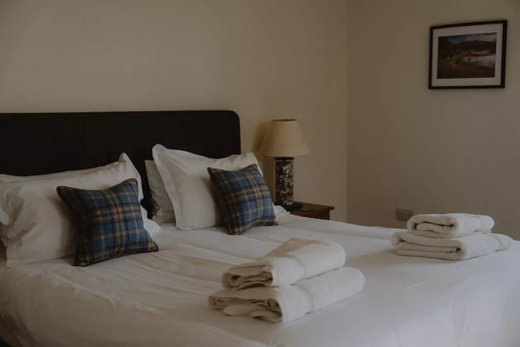 A bed dressed in white linen with tartan cushions and folded towels sits below a framed landscape print, a lamp lit on the bedside table.
