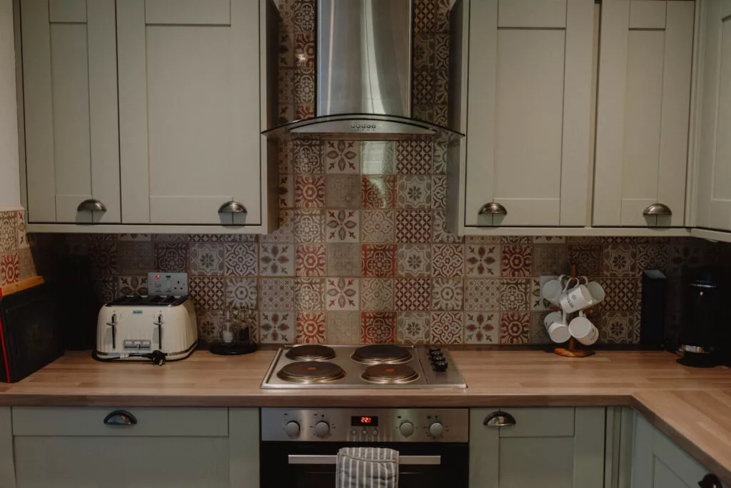 A pale green kitchen has a patterned tile splashback behind a stainless steel extractor hood and electric hob, a toaster and kettle on the counter.