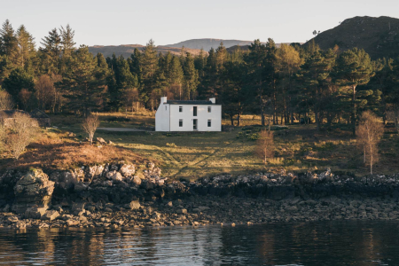 A white two-storey house stands on a grassy point above a rocky shoreline, pine trees rising behind it at sunset.