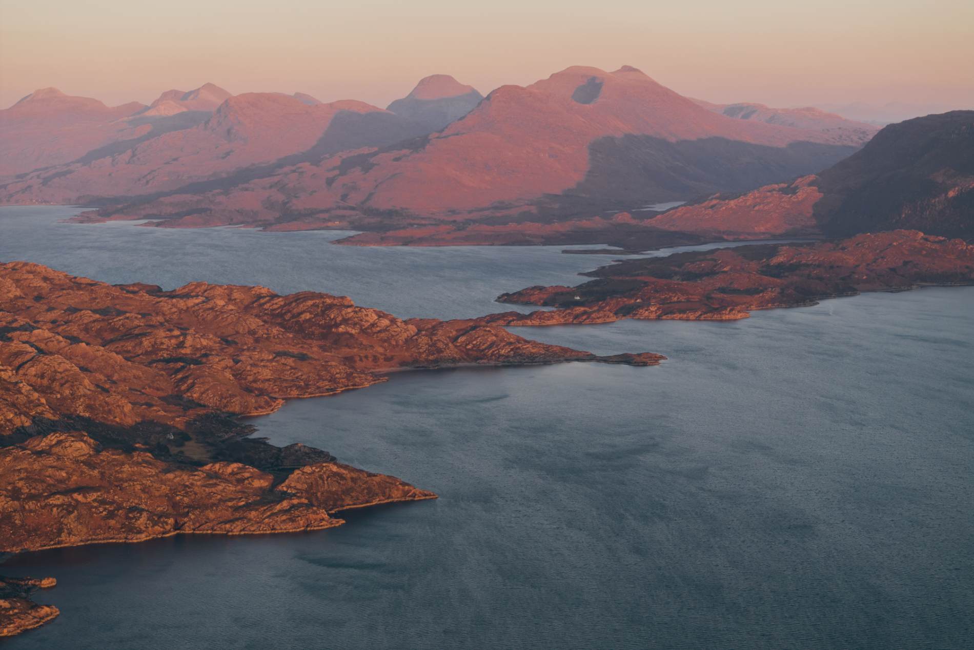 Mountain peaks catch the pink light of dawn above a loch and rugged coastline.