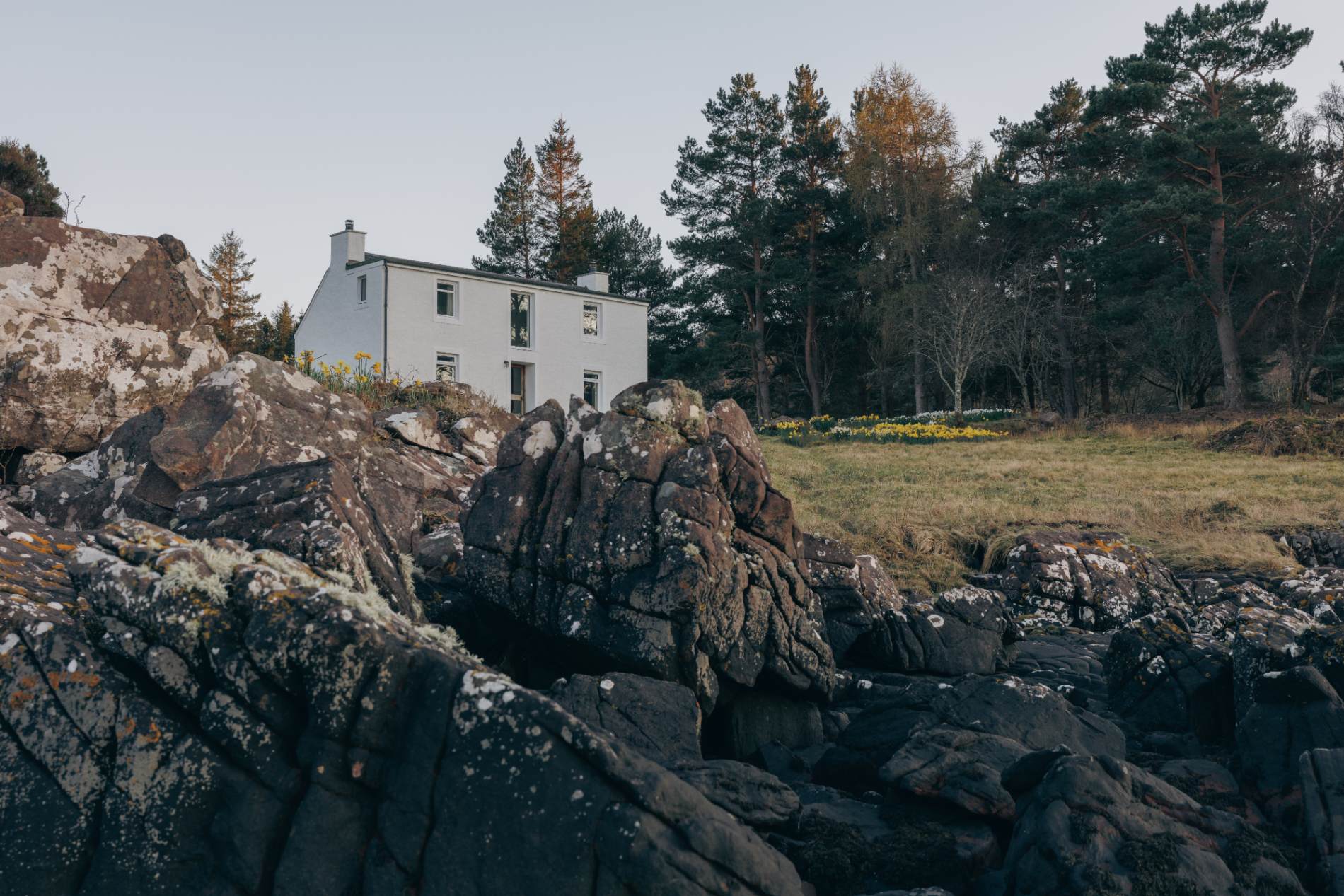 A white house is glimpsed beyond weathered coastal rocks, pine trees and autumn foliage framing the scene.