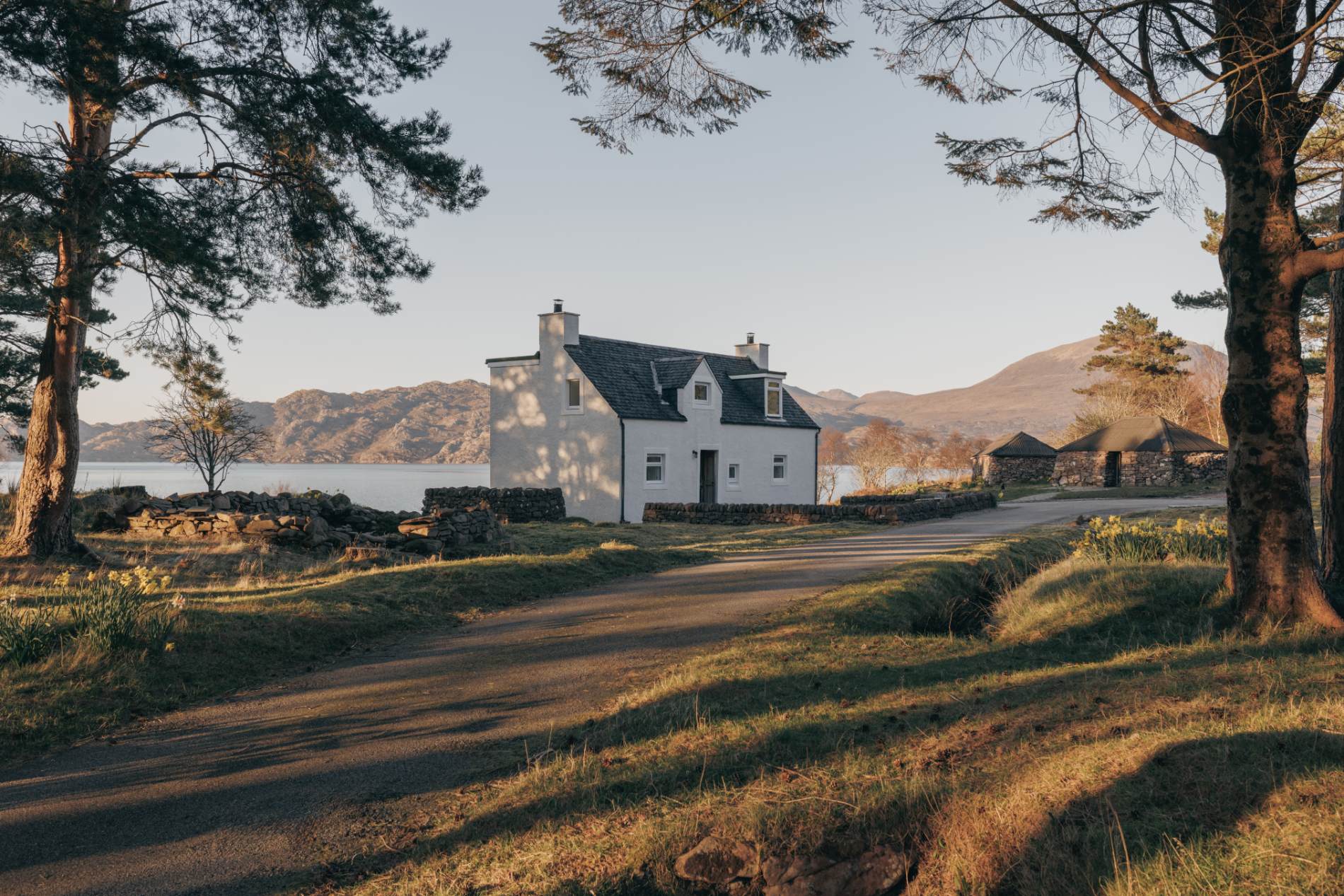 A white cottage with black-framed dormer windows sits above a loch shore, a gravel path curving past outbuildings behind.