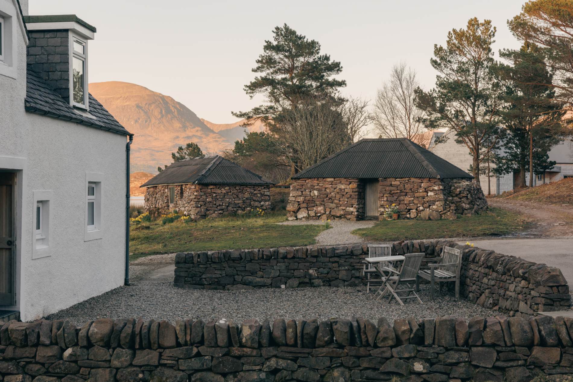 Stone bothies with dark slated roofs sit around a gravel courtyard, hills visible beyond a low stone wall with a table and chairs.
