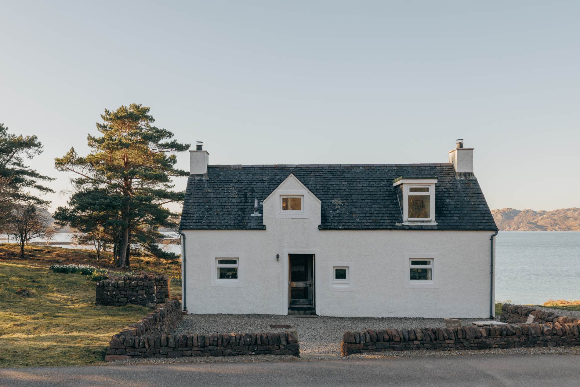 A white cottage with black-framed windows and twin chimneys stands beside a loch, a low stone wall and daffodils in the foreground.
