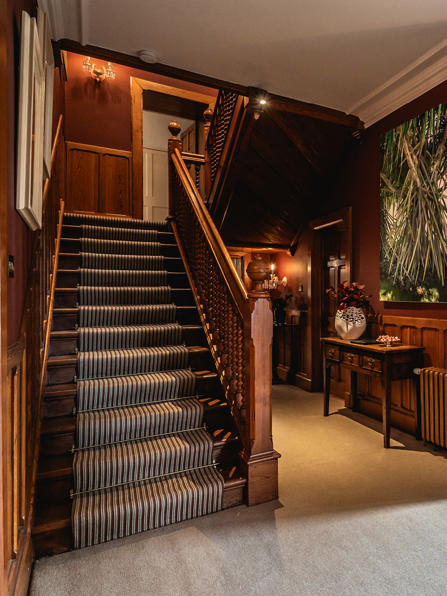 A striped stair runner climbs a wooden staircase beneath a carved balustrade, a console table with flowers visible in the hallway below.