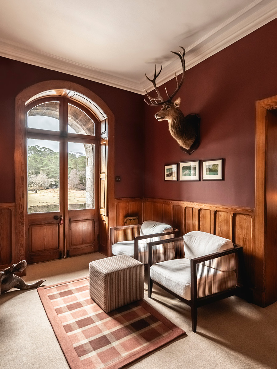 A mounted stag's head hangs above framed prints on a deep red wall, wicker armchairs and a striped stool arranged before an arched glazed door.