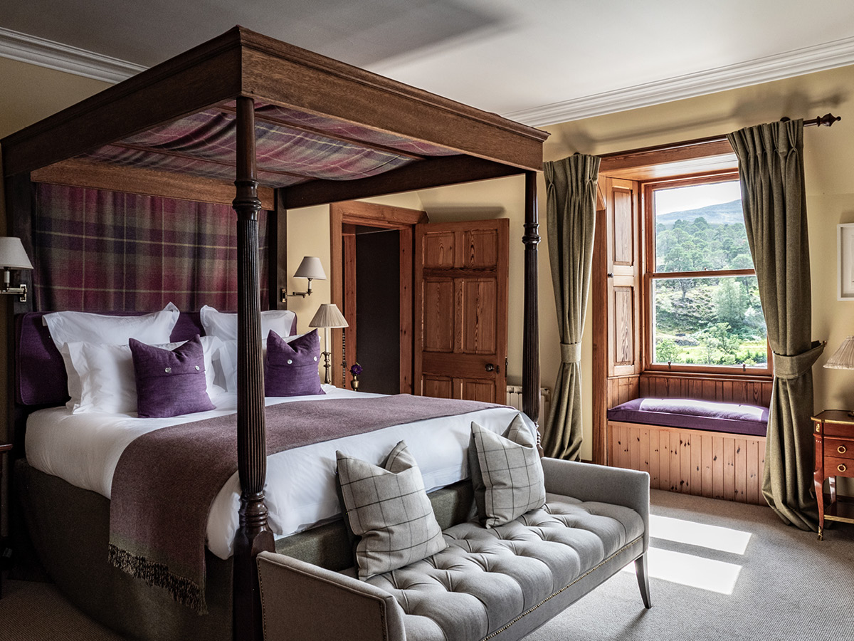 A four-poster bed with a tartan canopy and purple cushions sits before a curtained window seat, a grey sofa at its foot.