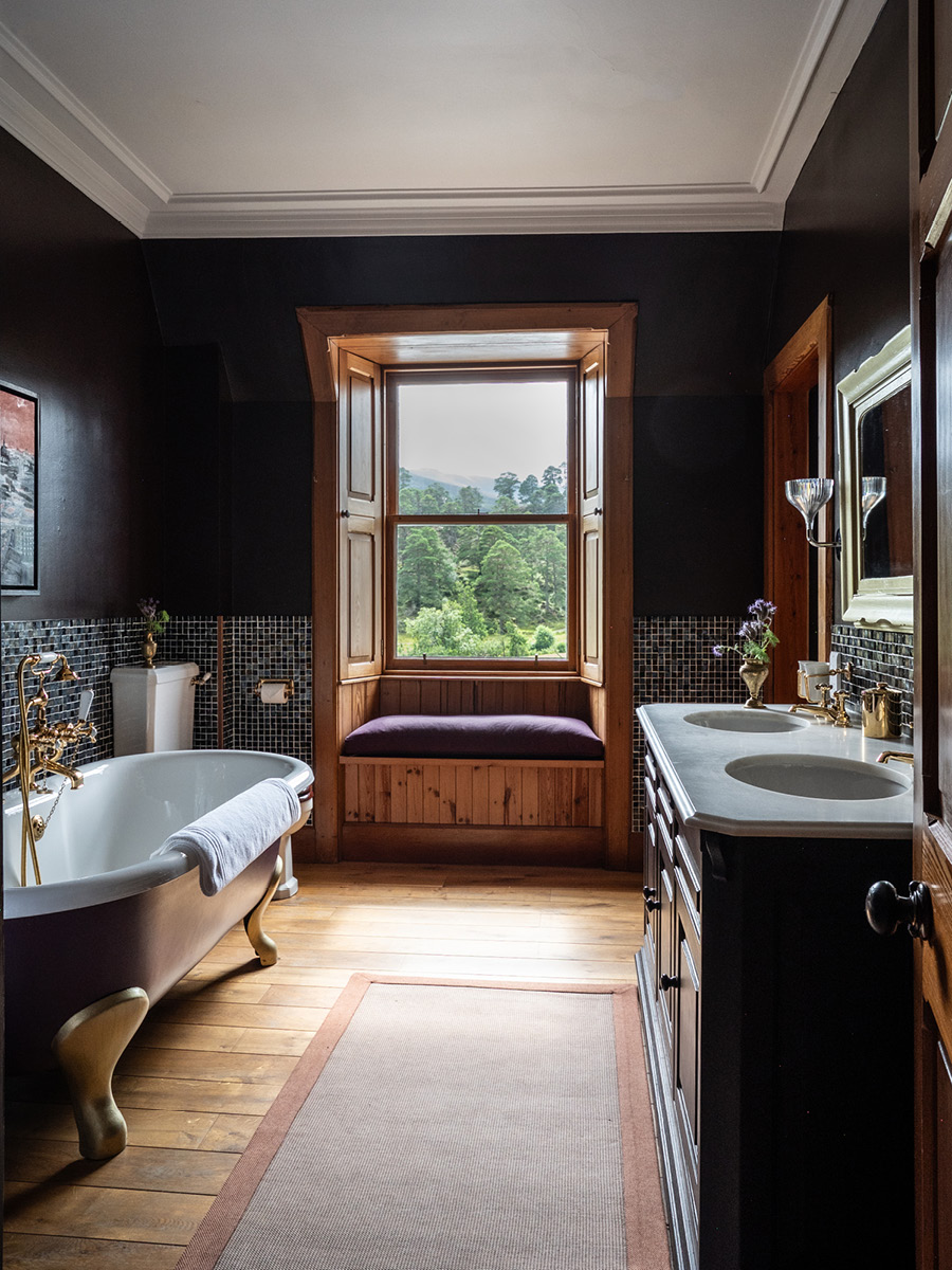 A roll-top bath stands beside a window seat with a purple cushion, twin basins and a mirror reflecting the room in dark panelling.
