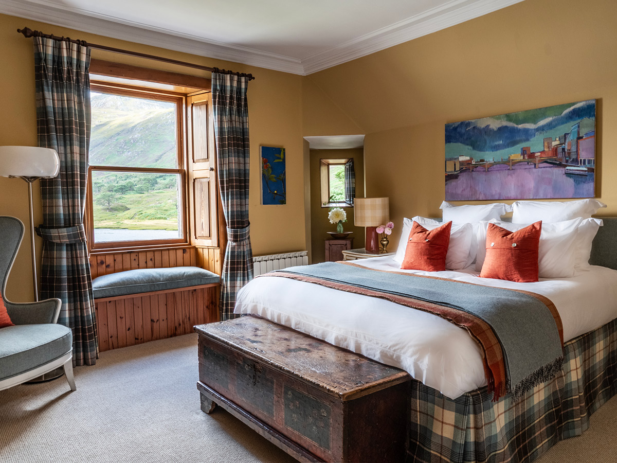 A bed with tartan throw and orange cushions sits below a mountain-themed painting, a window seat and wing-back chair completing the room.