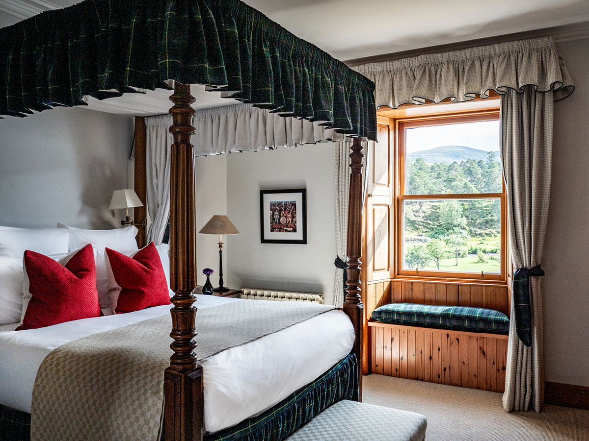 A four-poster bed with a tartan canopy and red cushions sits before a window seat looking onto hills and trees.