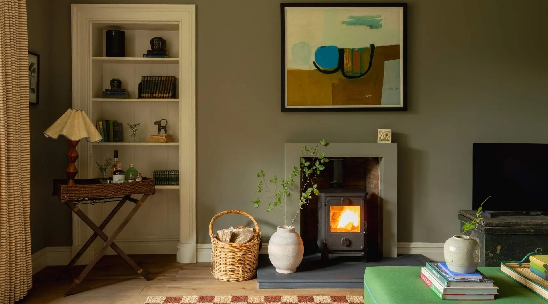 A wood burning stove alight in a pale stone surround, a basket of logs and a vase beside it, an alcove of shelved books above a side table with a lamp.