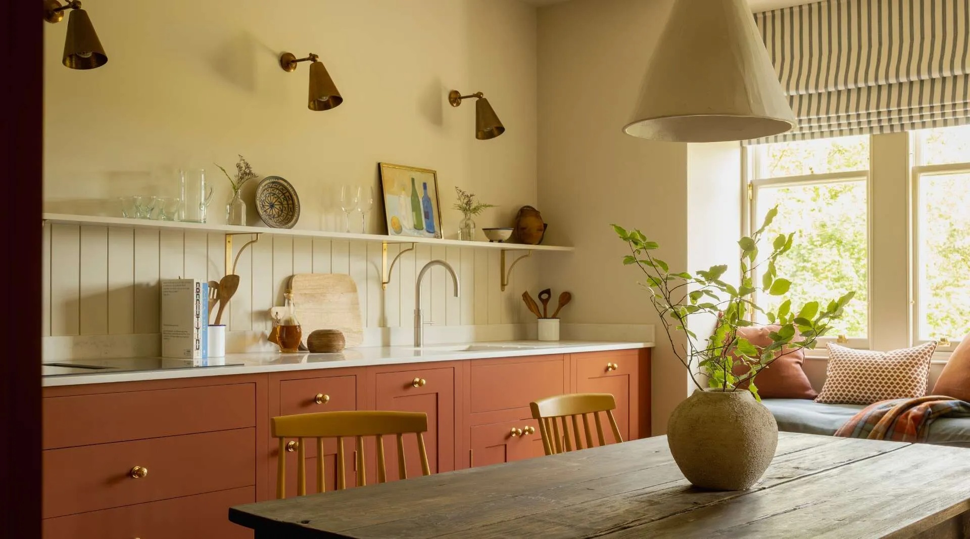 A kitchen with terracotta cabinetry and brass taps, a window seat with cushions and a vase of roses on the wooden table in front.