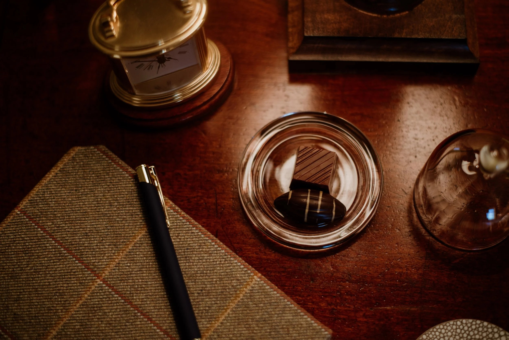 A leather notebook and pen rest beside a small dish of chocolates and a brass barometer on a polished wooden desk.