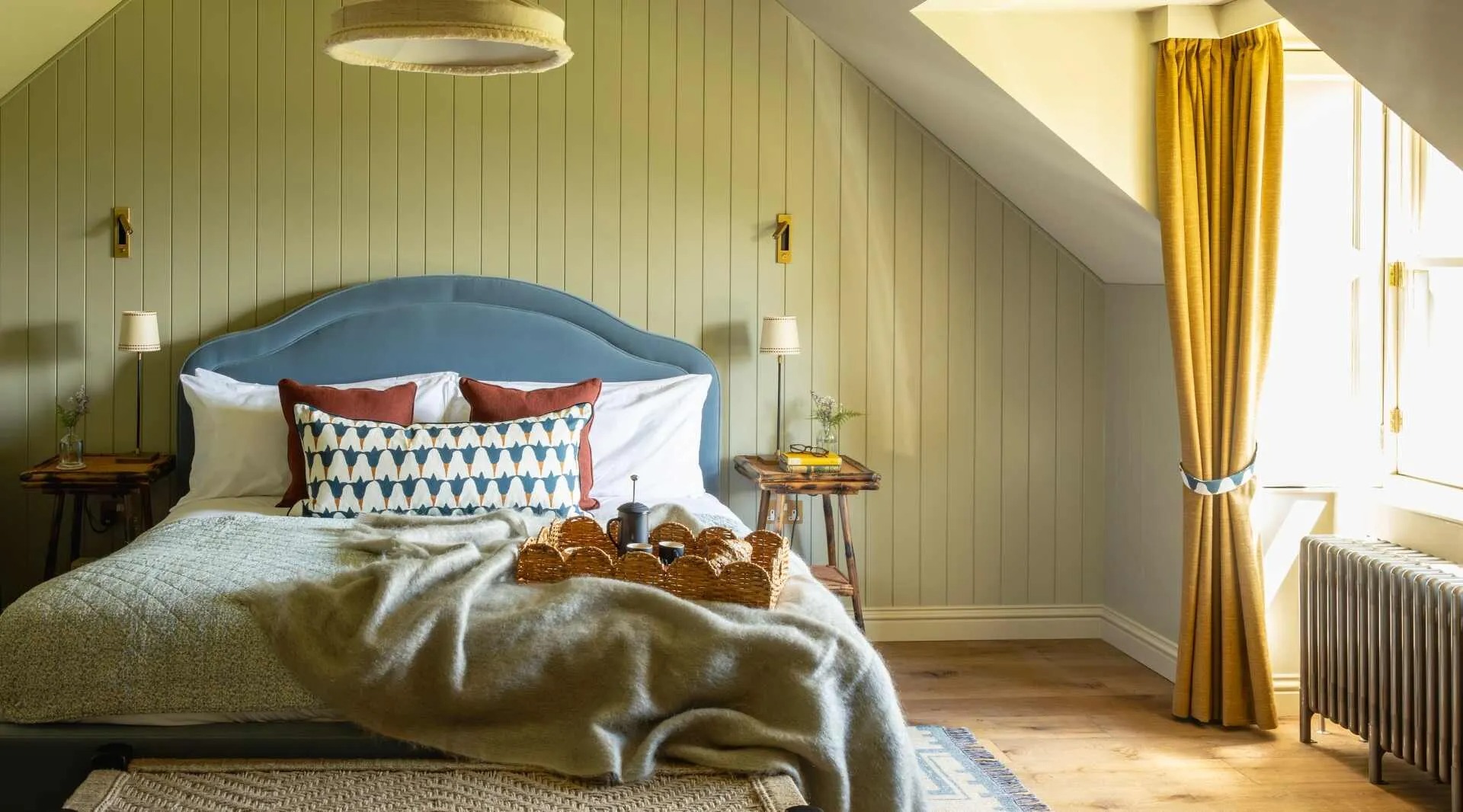 A bed with a blue upholstered headboard and patterned cushions sits under a sloped ceiling, a breakfast tray of pastries laid across the foot.