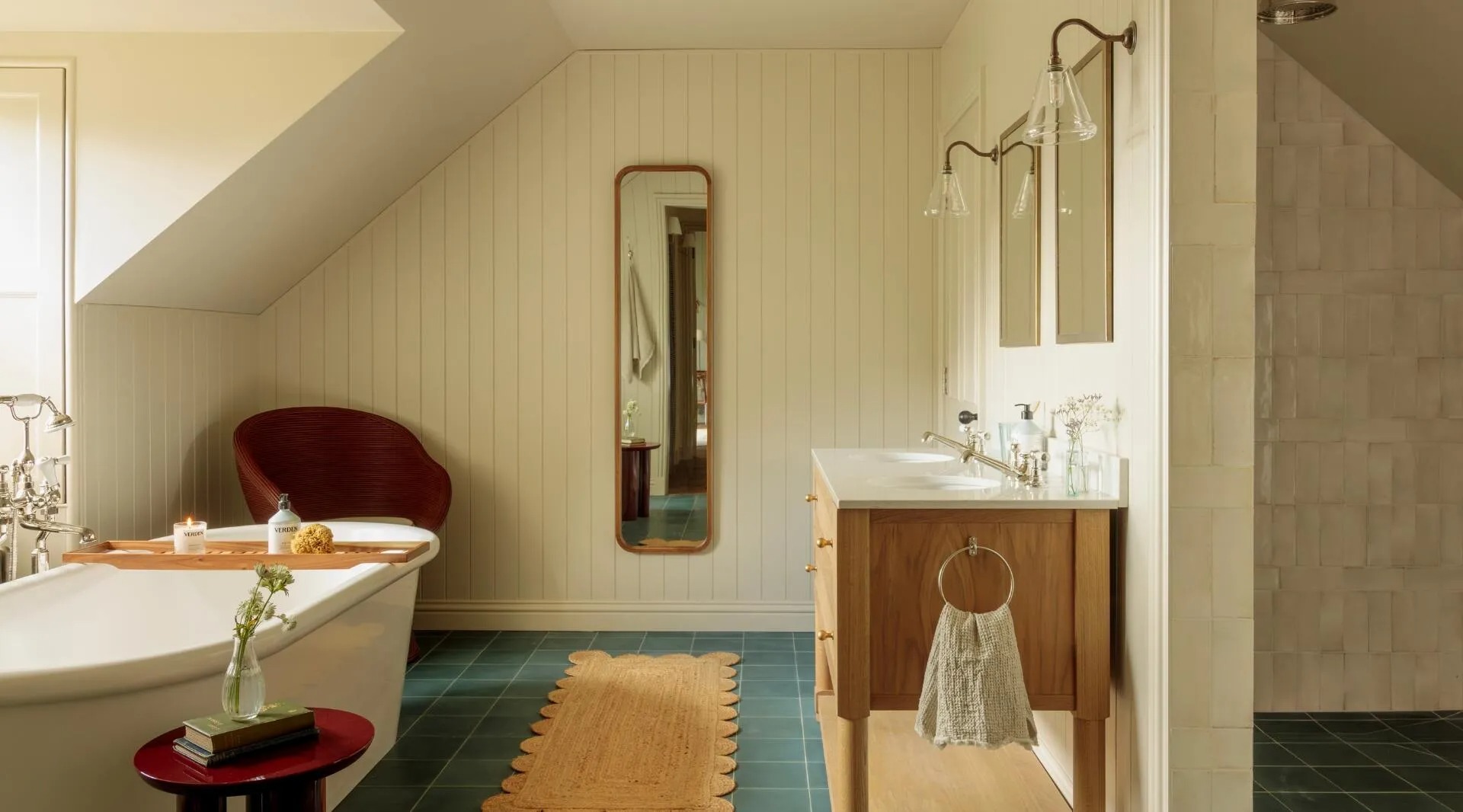 A freestanding bath with a red-lined interior sits beneath a sloped ceiling, a wooden vanity and shower area visible along the panelled wall.
