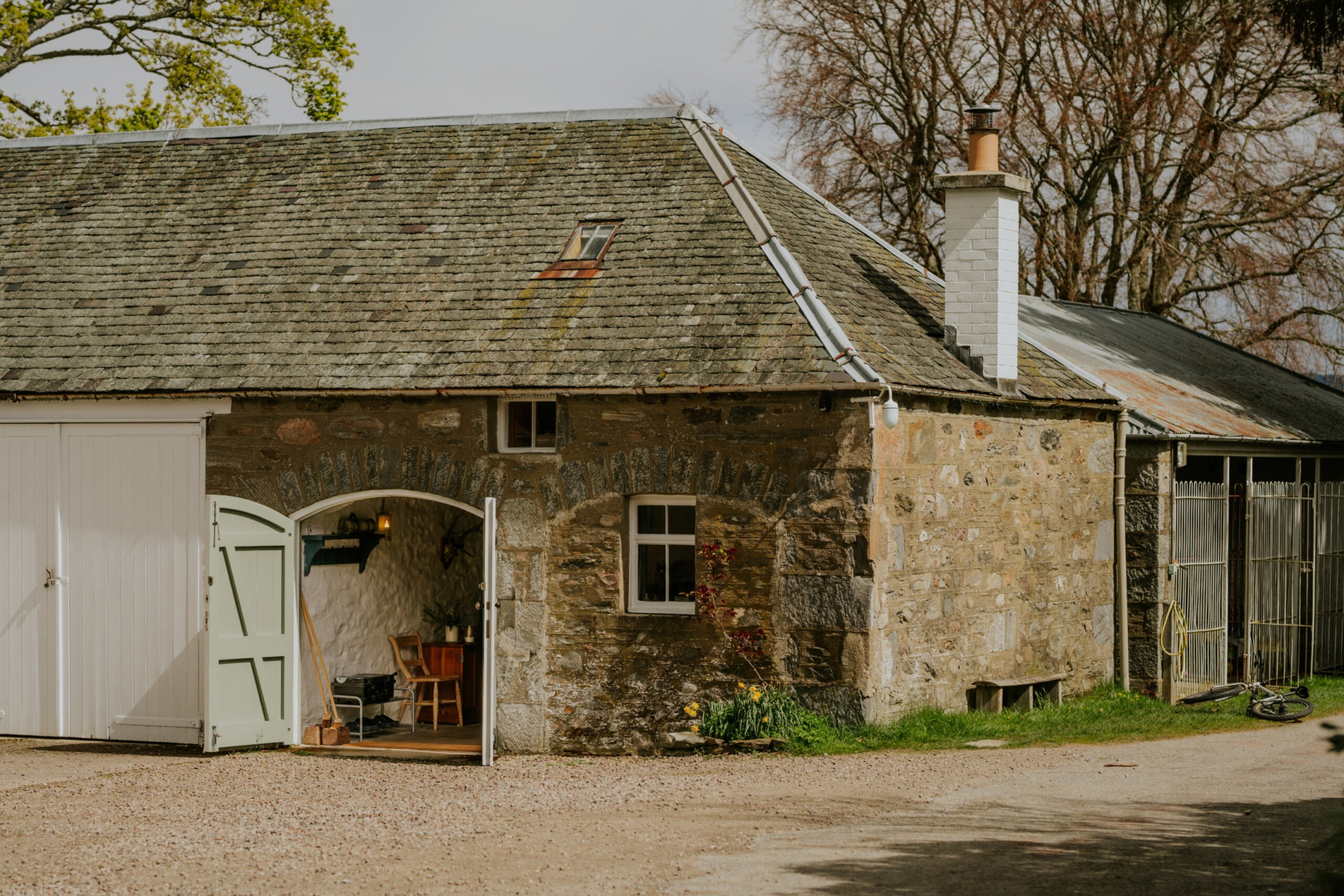 A stone steading with a slated roof and an arched green door stands beside a gravel courtyard, daffodils growing at its base.