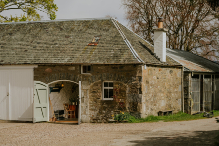 A stone steading with a slated roof and an arched green door stands beside a gravel courtyard, daffodils growing at its base.
