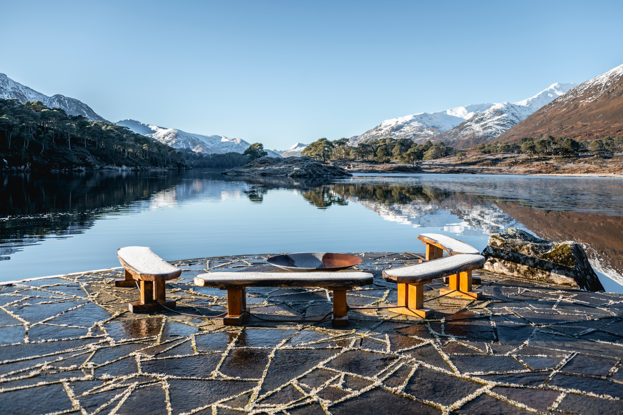 A stone fire bowl stands between two curved wooden benches on a slate terrace beside a still loch, snow-capped mountains reflected in the water.