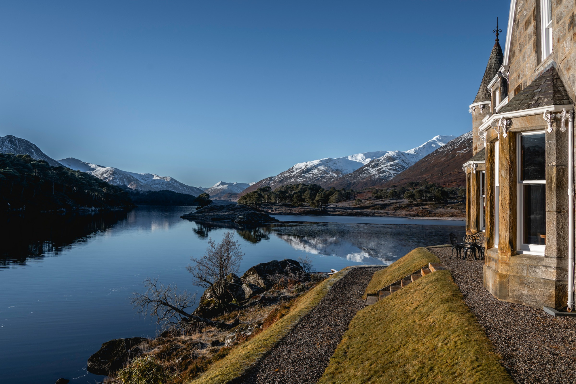 A turreted stone house catches the morning sun beside a still loch, snow-capped mountains reflected in the water.