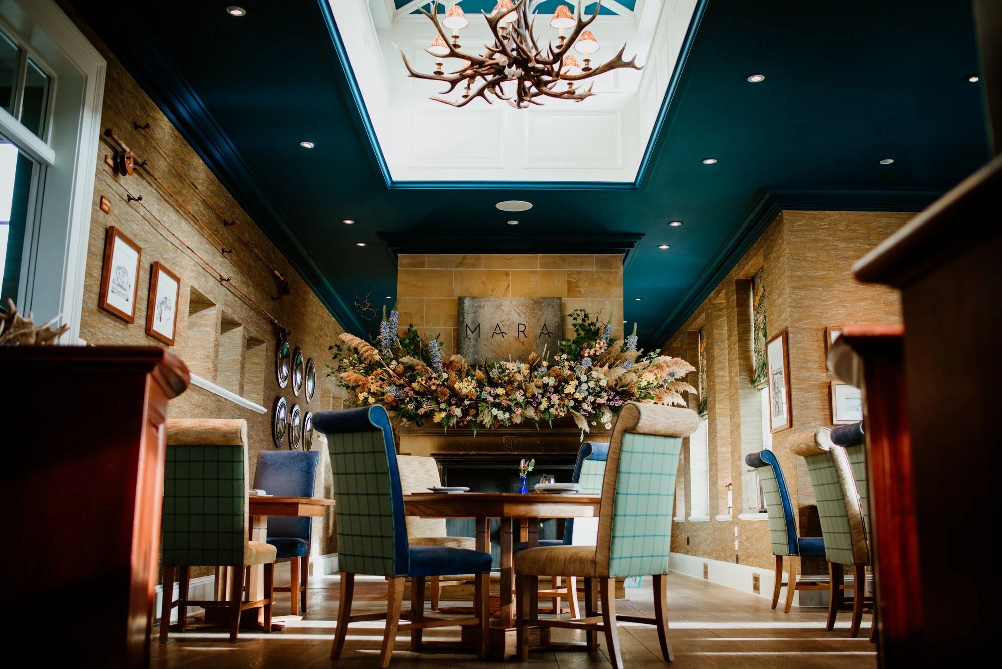 Wing-back chairs in check fabric surround tables beneath a domed skylight, a floral display and antler chandelier above the mantel.
