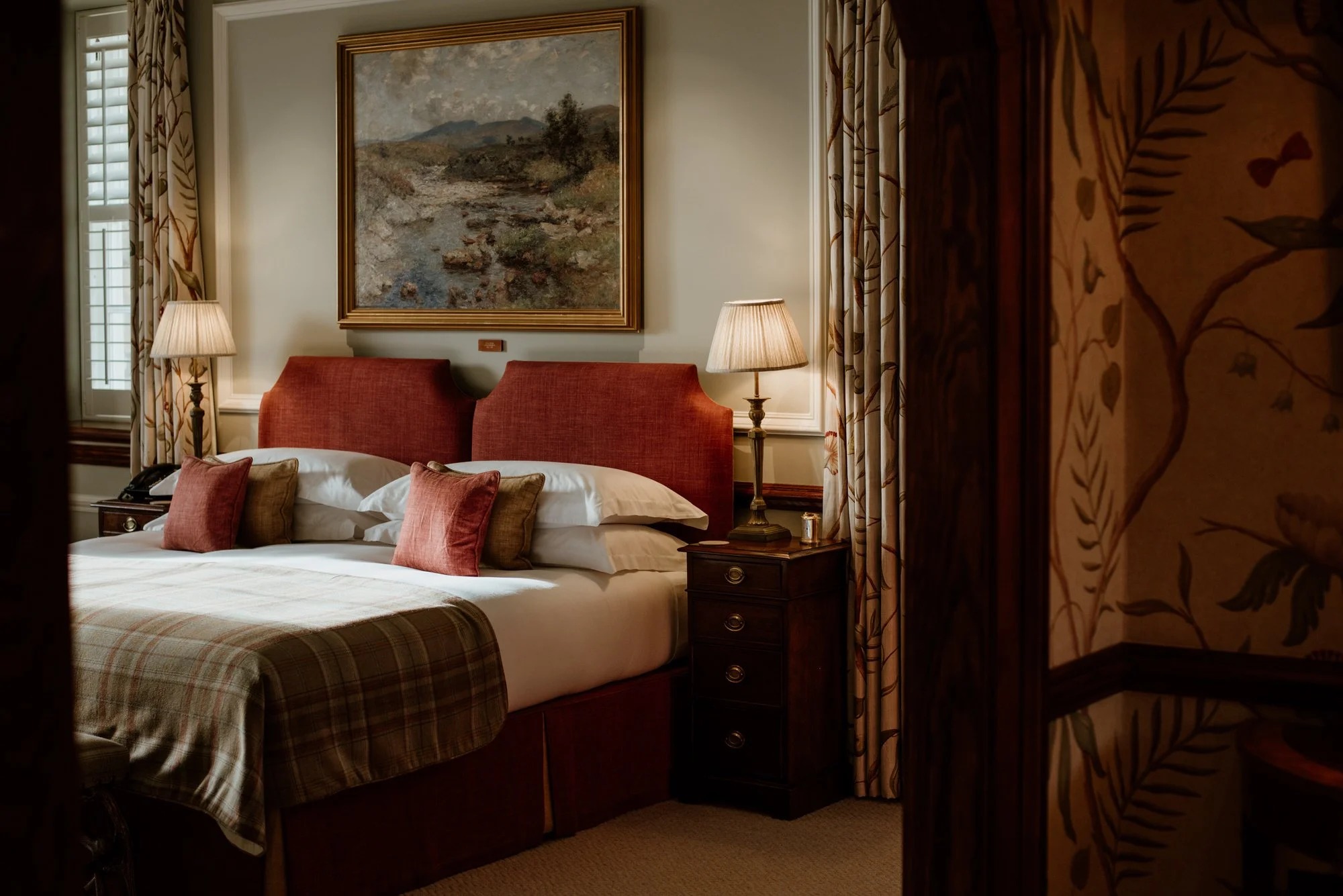 A bed with a red upholstered headboard and check throw sits beneath a landscape painting, patterned curtains framing the doorway.