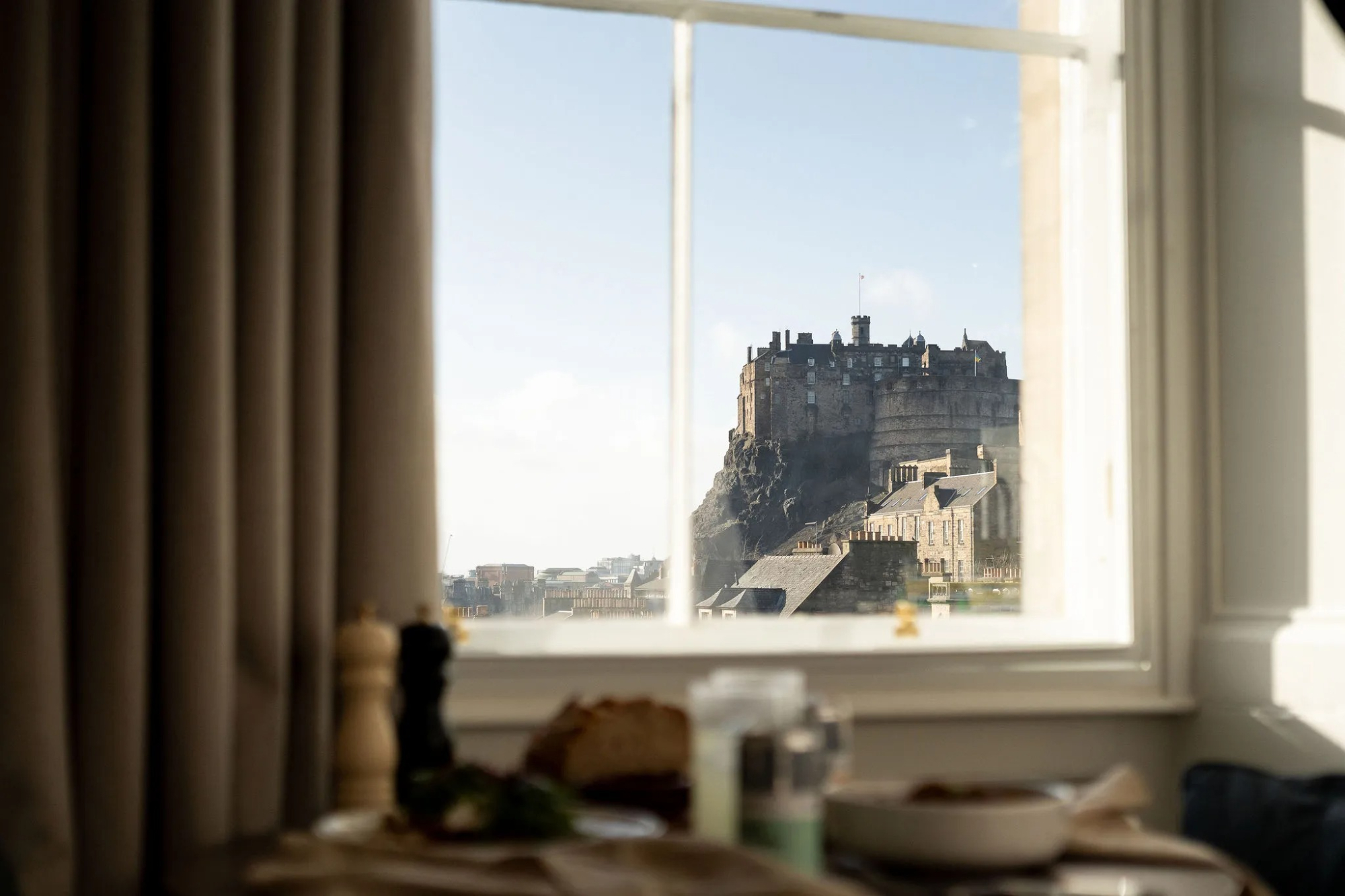 A castle on a rocky outcrop is framed through a sash window, a pepper grinder and plates blurred on the table below.