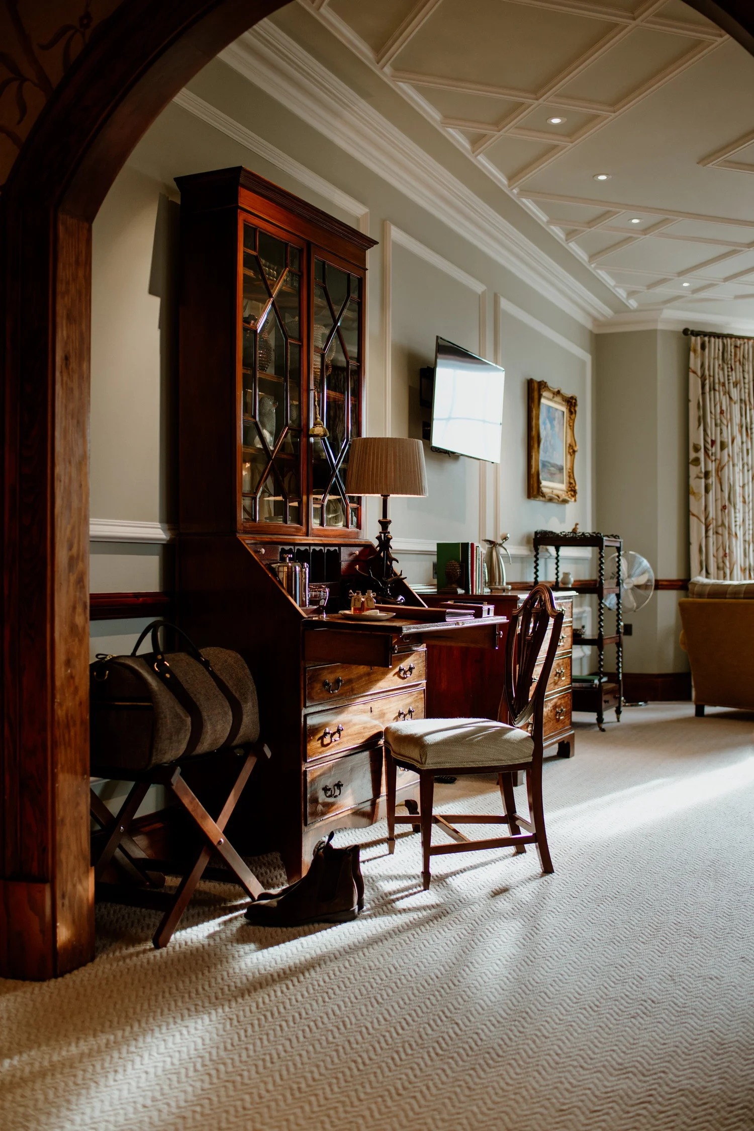 An antique writing desk with a glass-fronted bookcase stands beneath a coffered ceiling, a mounted television and armchair to the side.