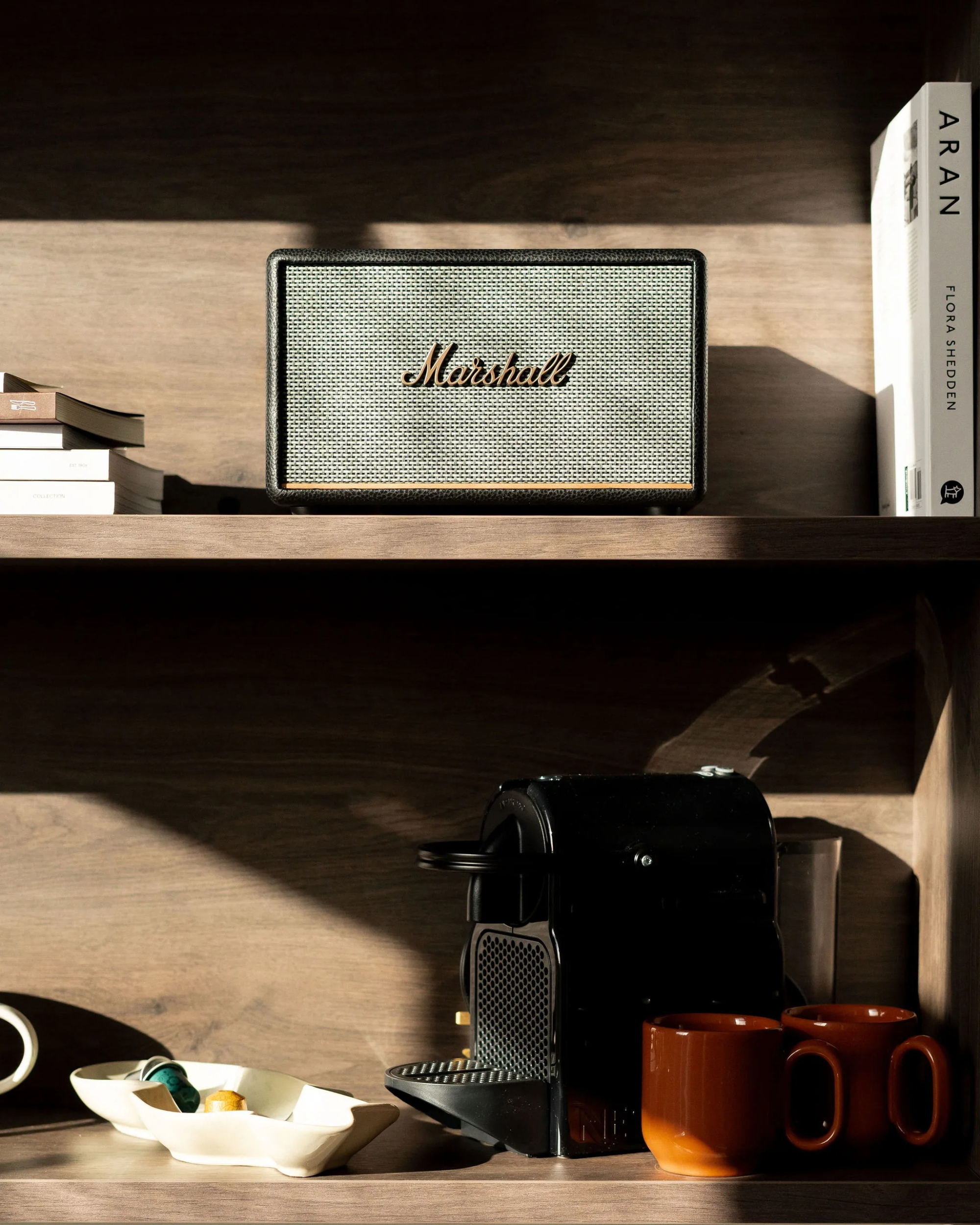 A speaker sits on a wooden shelf beside a stack of books including "Aran" by Flora Shedden, a coffee machine and mugs on the shelf below.