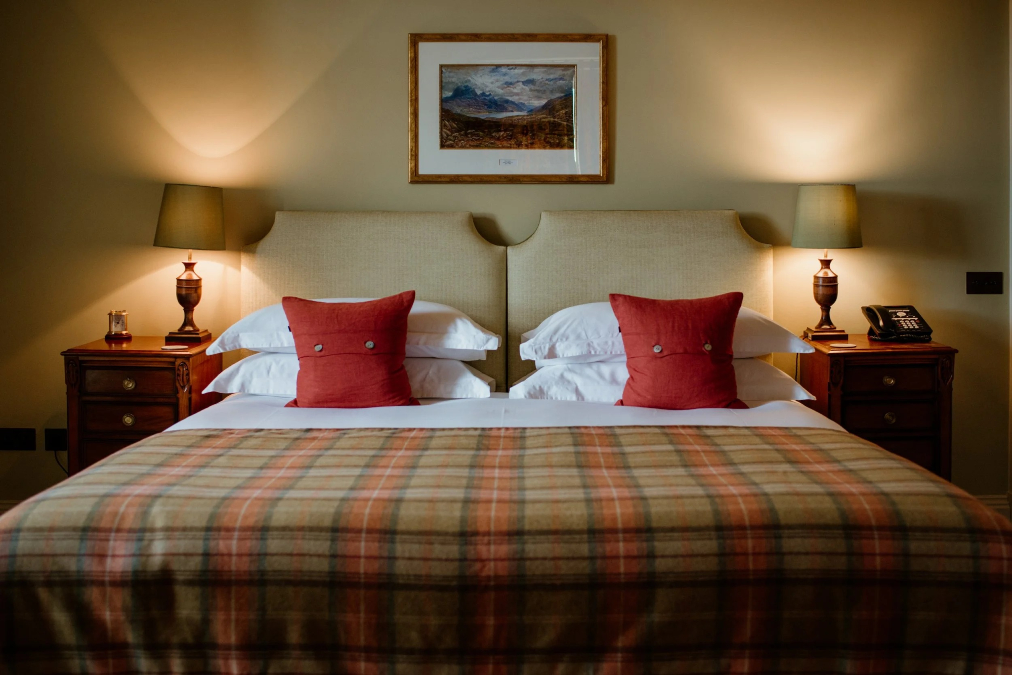 A bed with red cushions and a plaid throw sits between two lit lamps, a framed landscape painting above the headboard.
