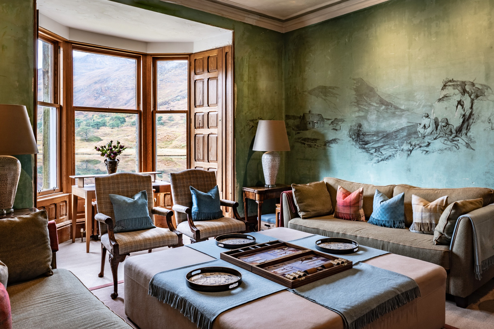 A backgammon set stands open on an ottoman before a green-walled sitting room with a bay window overlooking hills, a mural of a landscape painted on the wall.