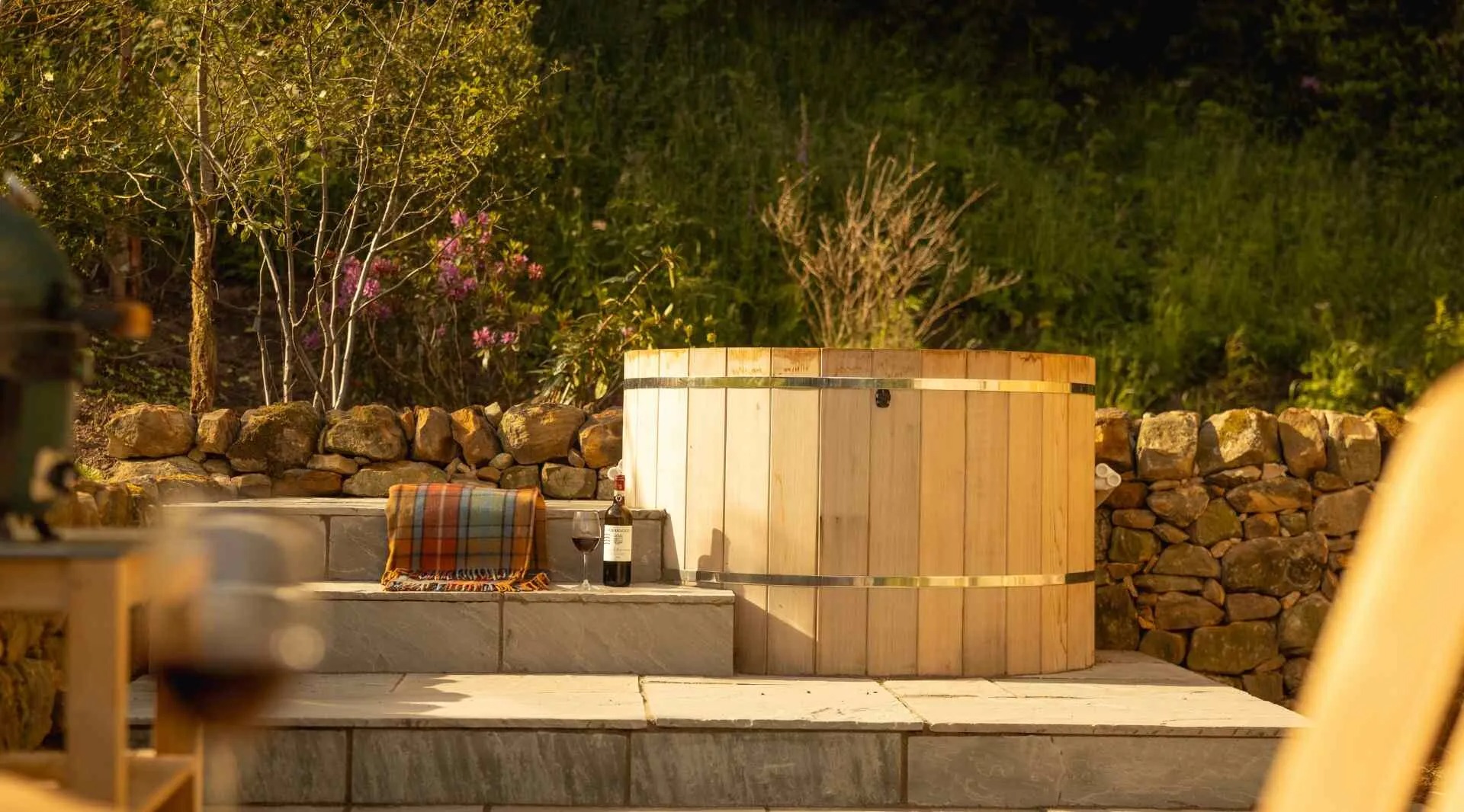 A wood-fired hot tub with steps sits on a stone patio backed by a dry stone wall and flowering shrubs.