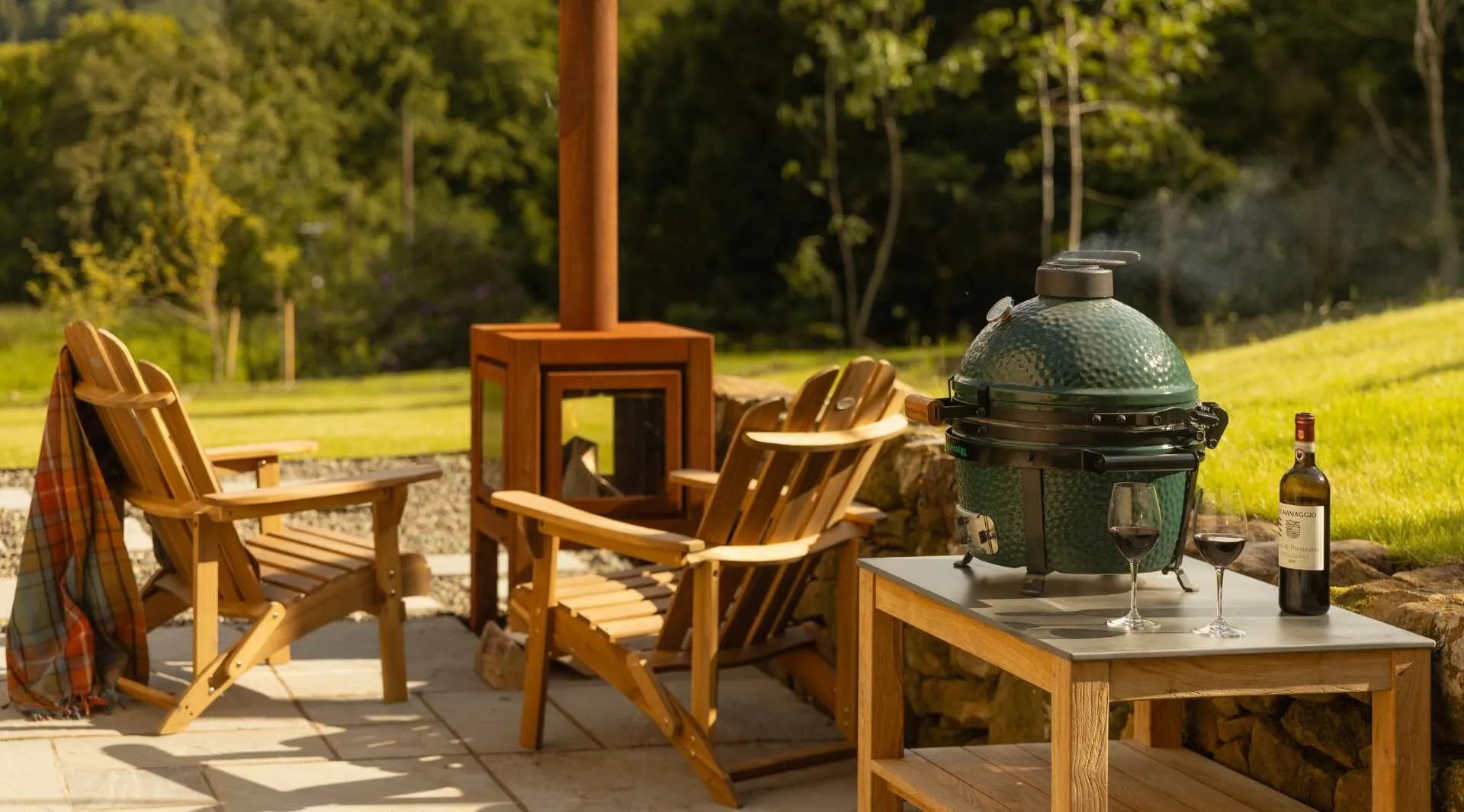 Wooden chairs and a wood-fired stove surround a table set with a kamado barbecue, a bottle of wine and glasses, on a stone patio.