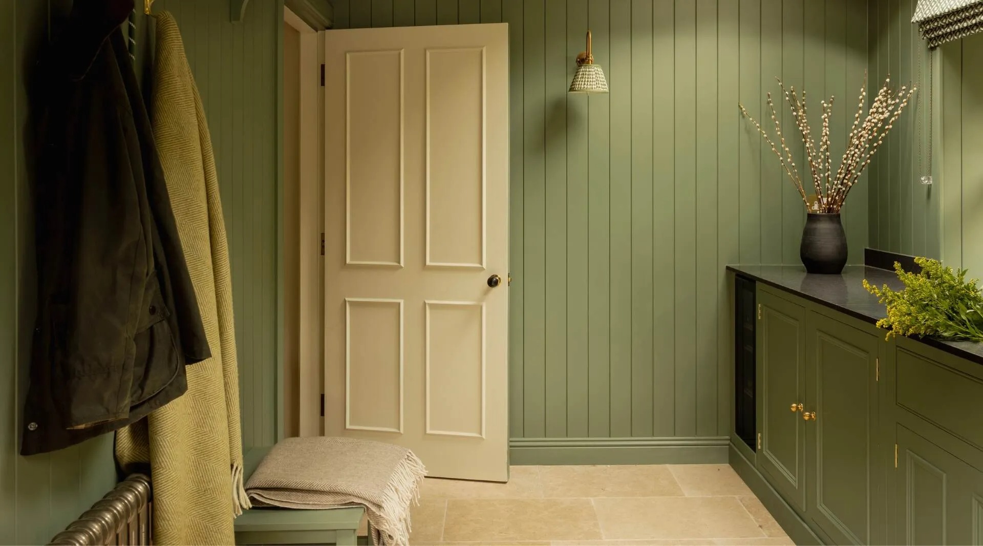 A boot room with sage green panelling, coats hung on pegs beside a bench and a dark cabinet topped with dried flowers.