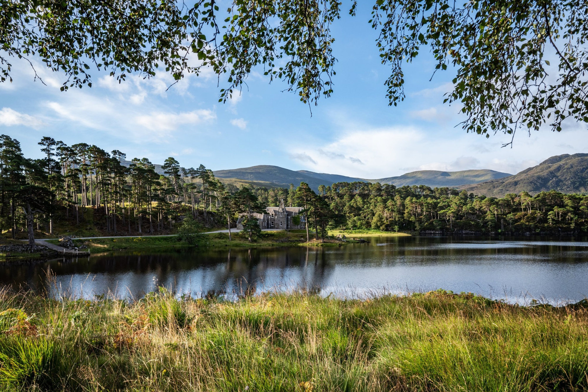 A turreted stone house sits among pine trees on a loch shore, hills rising behind under a clear sky, seen through overhanging birch leaves.