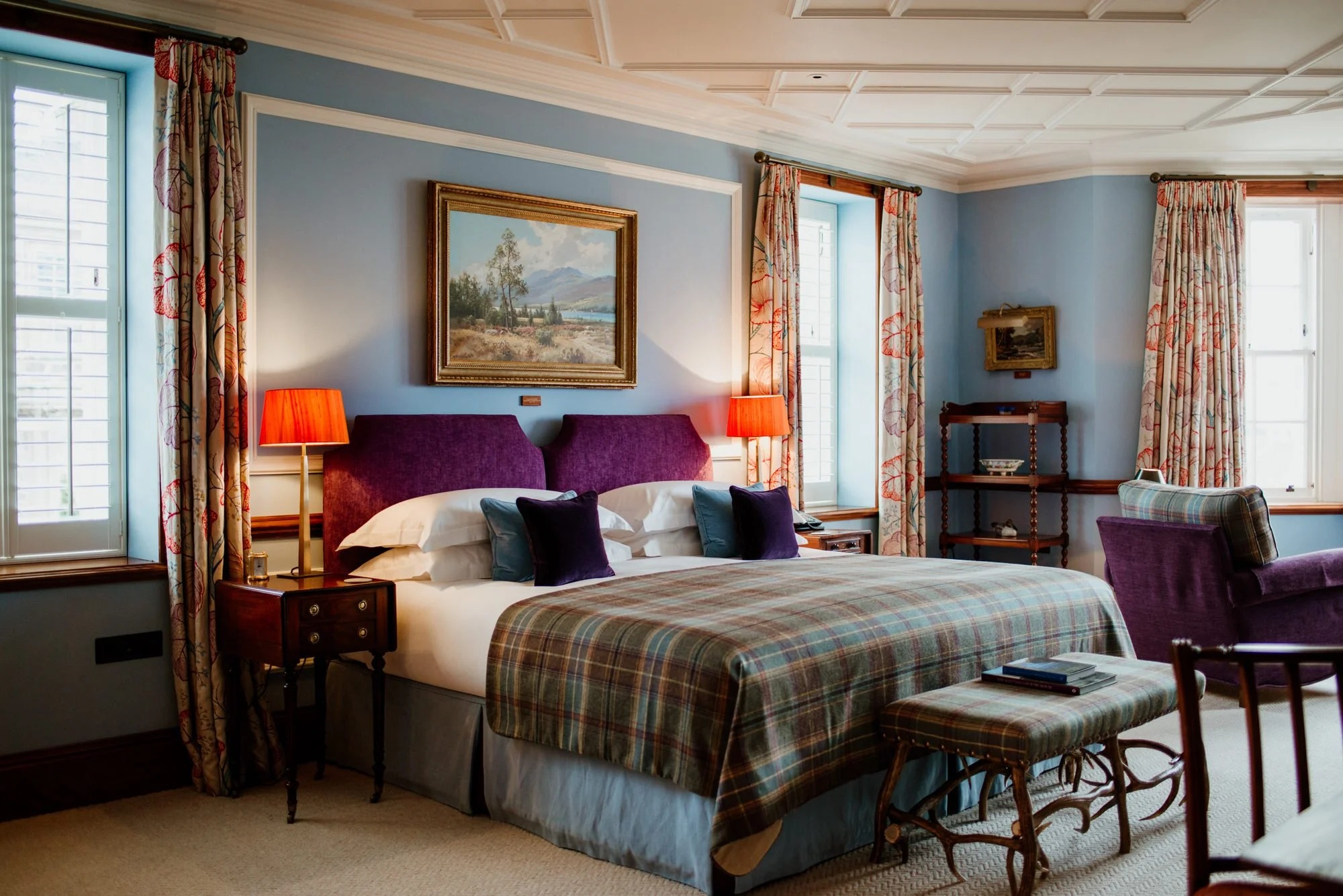 A bed with a purple upholstered headboard and tartan throw sits below a landscape painting, patterned curtains framing the windows either side.