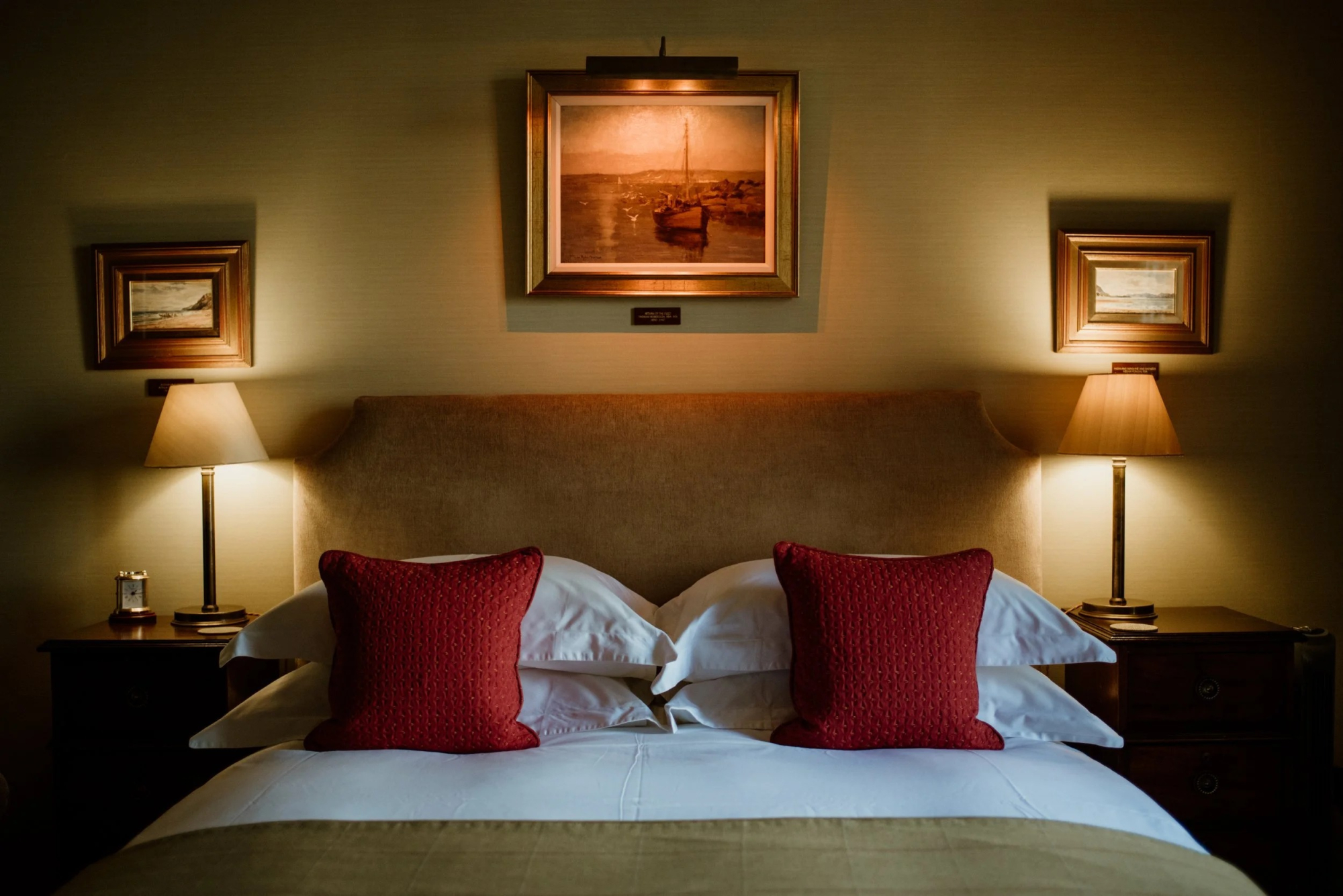 A bed with red cushions sits beneath a framed painting of a harbour scene, matching lamps lit on the bedside tables either side.